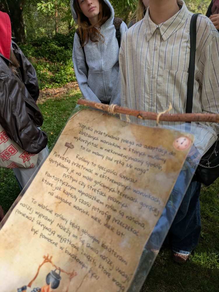 Zdjęcie przedstawia uczniów podczas warsztatów „Przyrodniczy survival” w Śląskim Ogrodzie Botanicznym w Mikołowie. Uczniowie zapoznają się z materiałami dotyczącymi zasad przetrwania i wykorzystania roślin w warunkach terenowych.